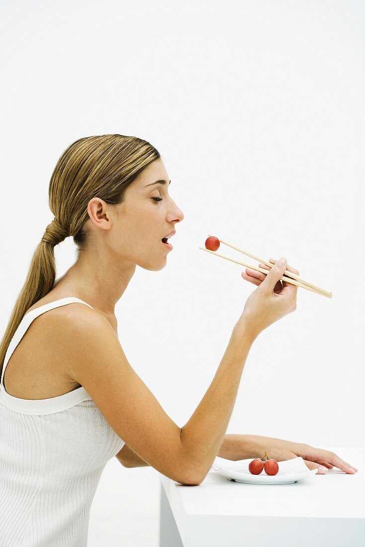 Woman eating red grape with chopsticks, eyes closed, side view