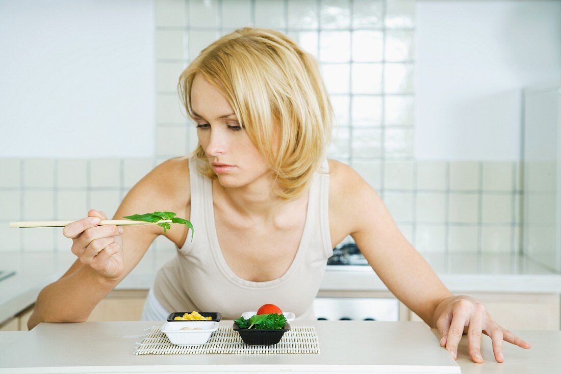Young woman leaning over kitchen counter, picking up herbs with chopsticks