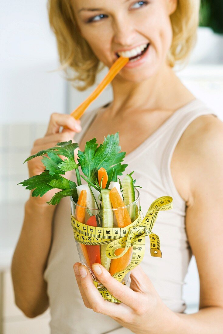 Frau beisst in Möhre und hält ein Glas Gemüsesticks mit Massband