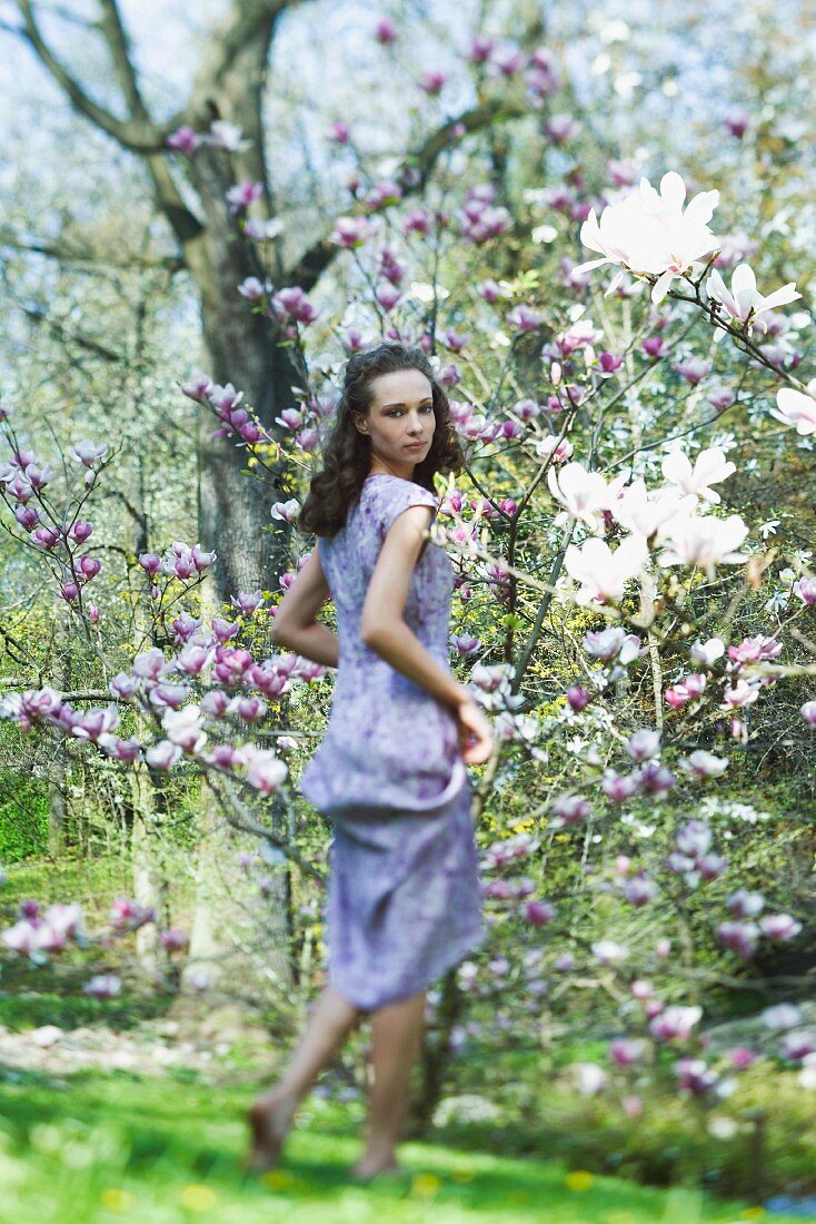 Young woman in dress walking beside flowering tree, looking over shoulder at camera