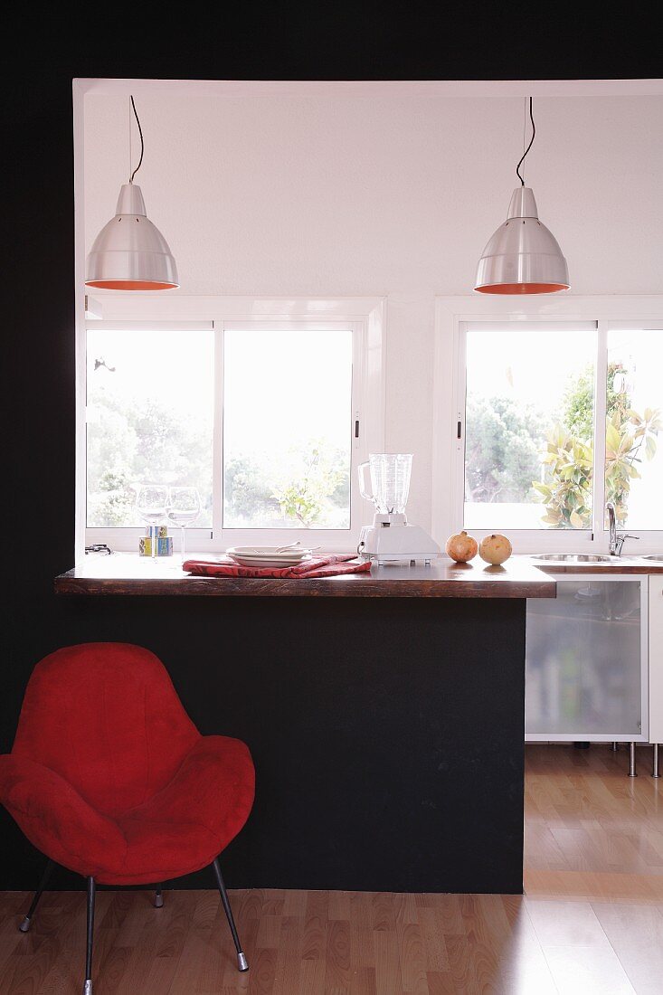 Roter Sessel vor schwarzer Wand mit Ausschnitt und eingebauter Esstheke und Blick in offene Küche im Designerstil