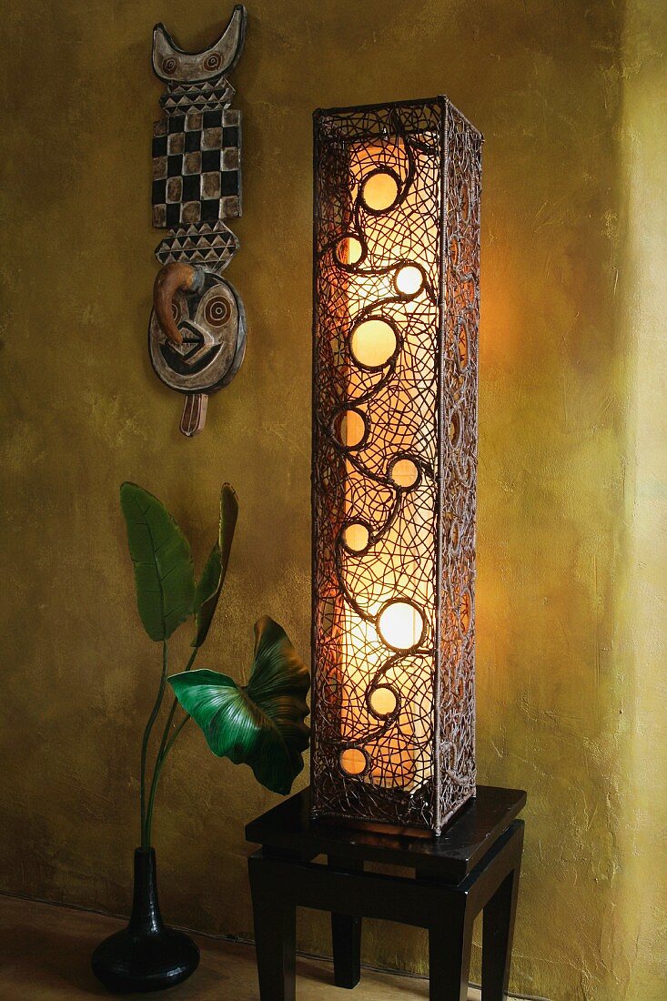 Stehlampe auf Hocker aus dunklem Holz und afrikanische Maske an getönter Wand