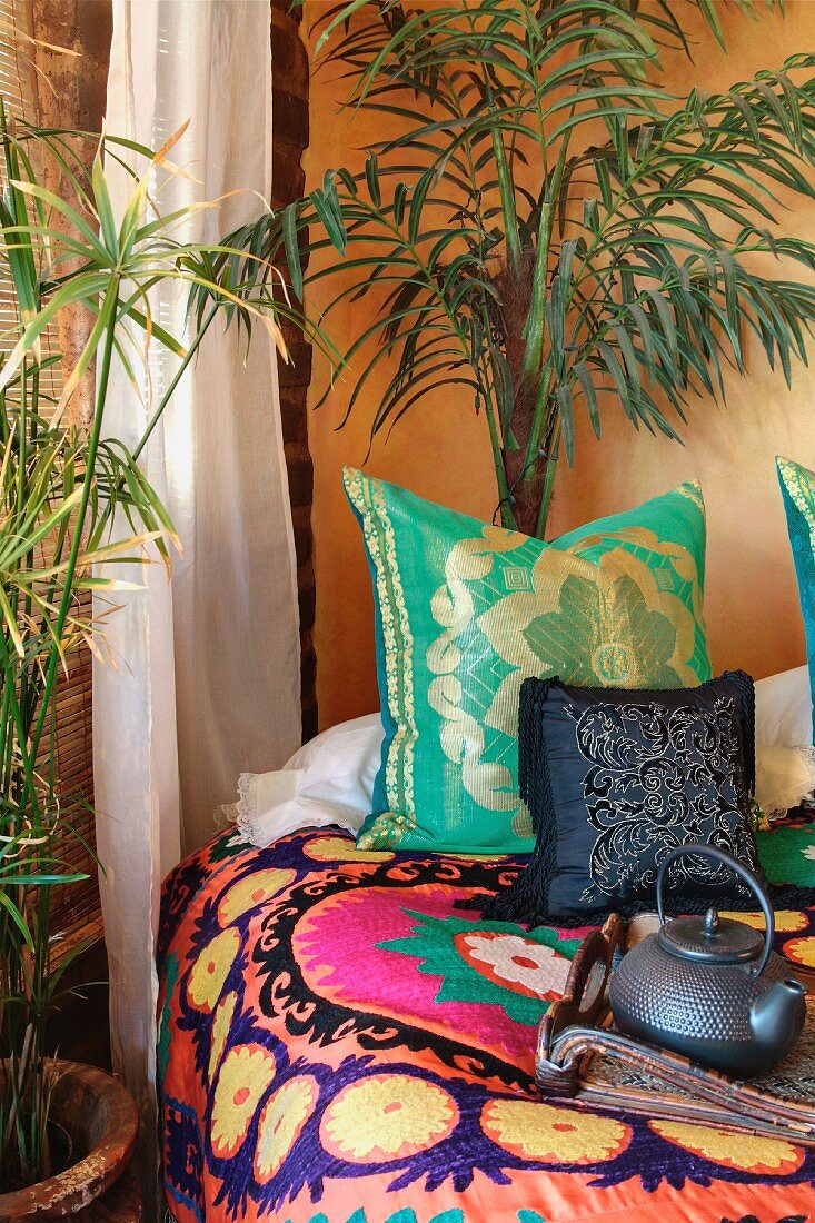 Schlafzimmerecke in afrikanischem Stil - Palmen neben Bett mit bunten Kissen und folkloristischer Tagesdecke