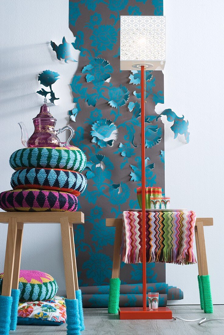 African cushions on wooden stools with knitted socks in front of floral wallpaper with some flowers cut and folded out