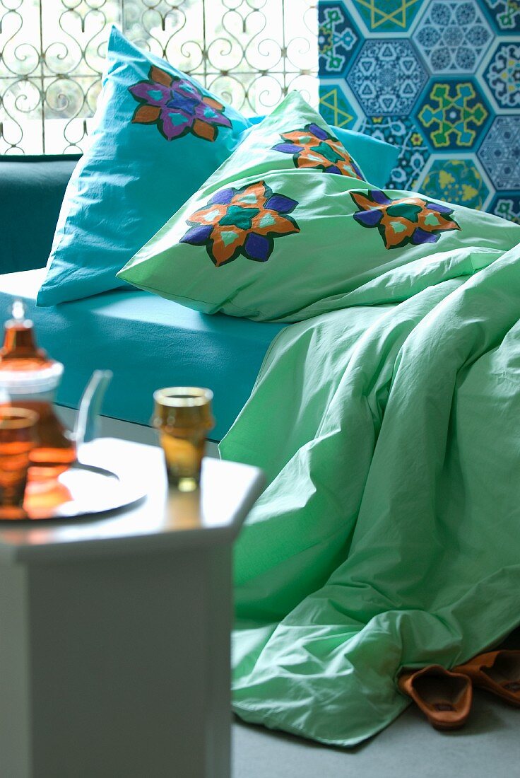 Tea set on side table in front of bed with colourful cushions and bedspread against wall with Oriental patterned tiles