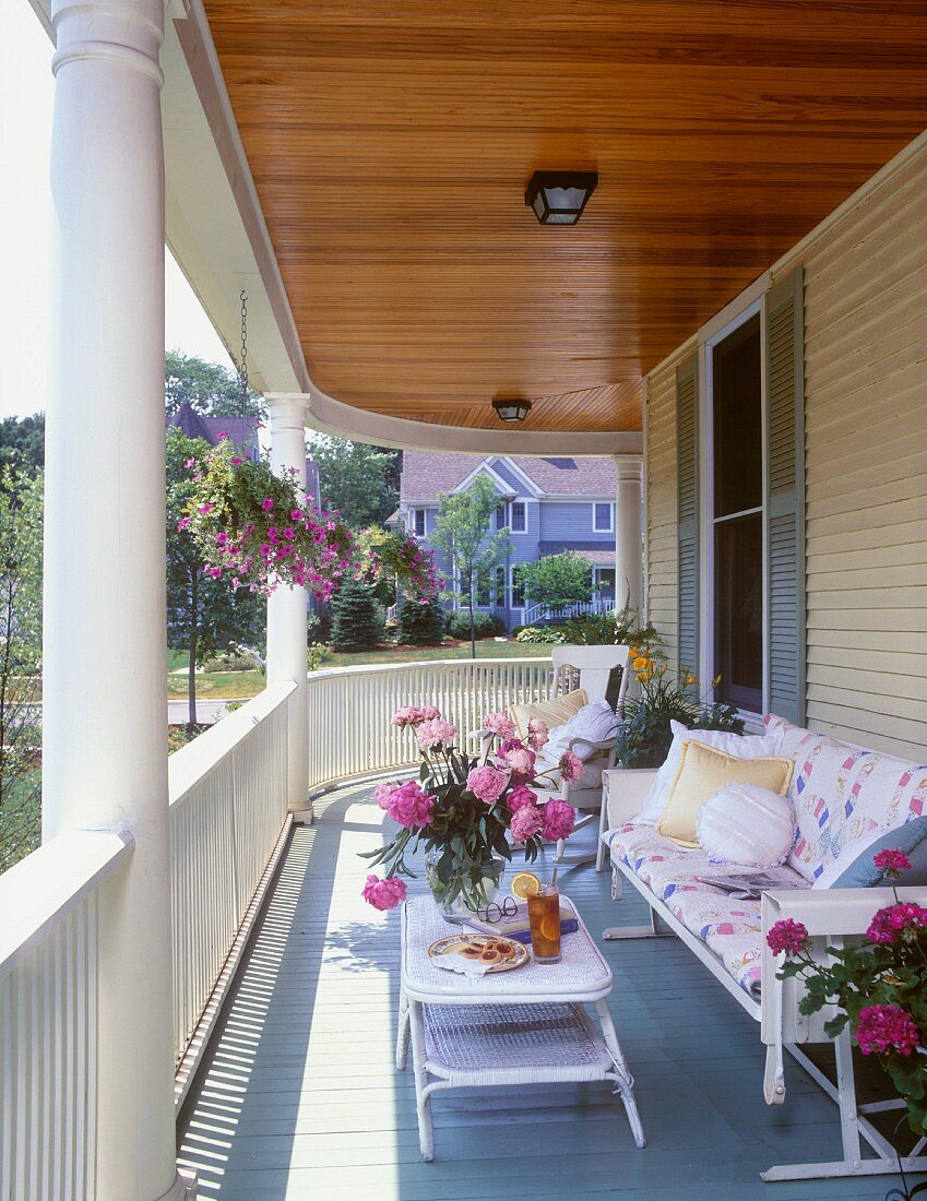 Geschwungener, sommerlicher Balkon eines Hauses mit Holzfassade mit bequemen Balkonmöbeln, Blumentöpfen und üppigem Pfingstrosenstrauss