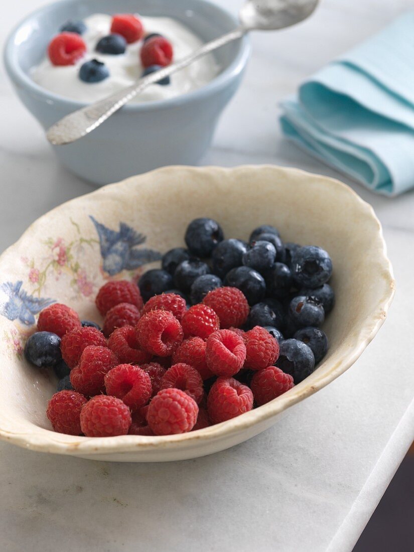 A Bowl of Raspberries and Blueberries; Bowl of Yogurt with Berries in Background