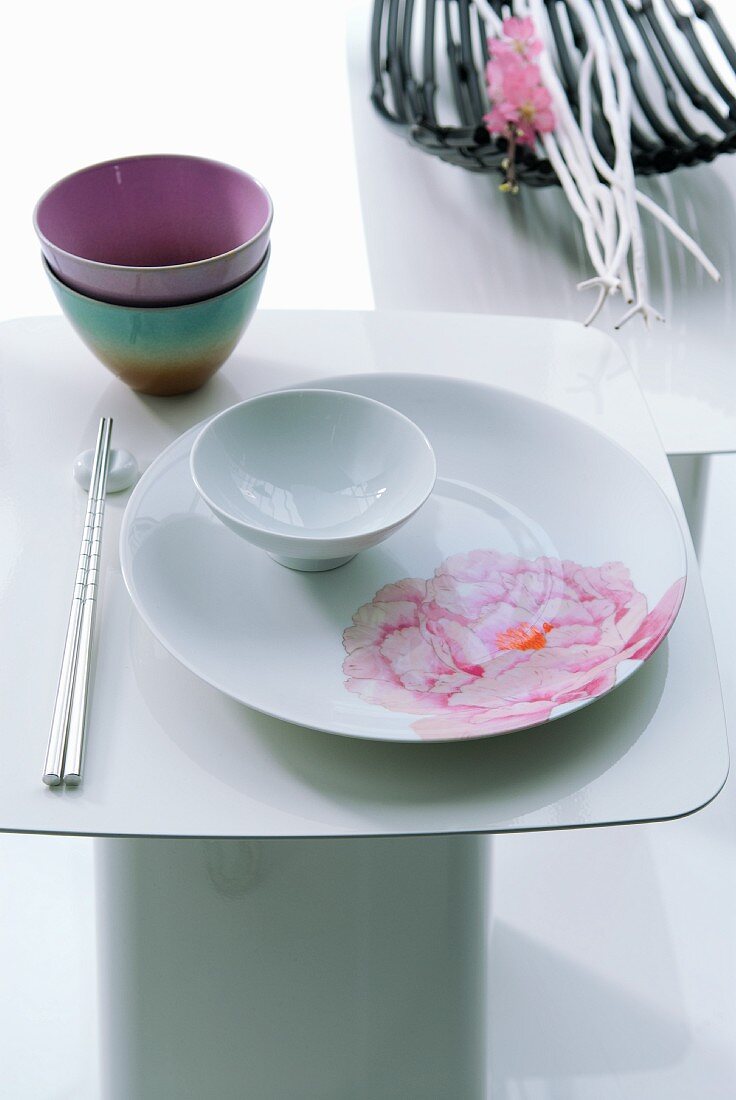 Japanese place setting on white side table with decorative bowl of twigs and blossom in background