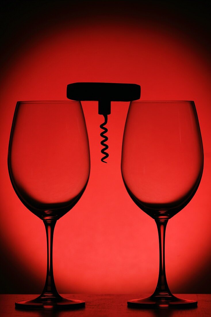 Zwei leere Weingläser und ein Korkenzieher vor roter Hintergrund