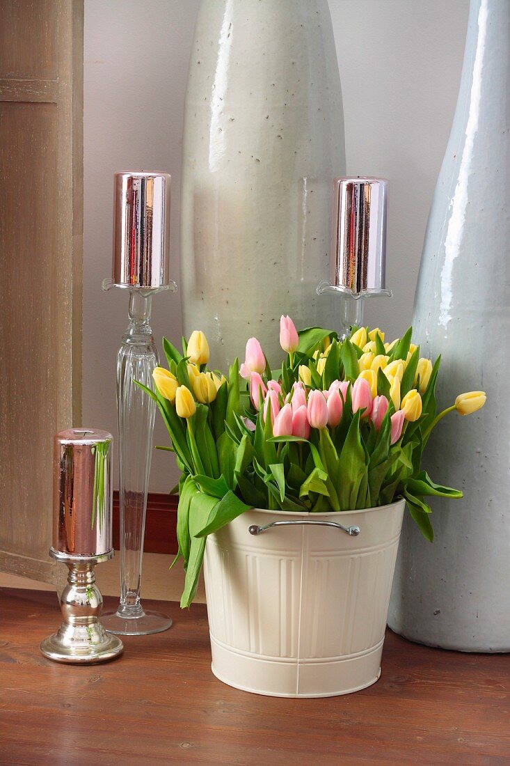 Yellow and pink tulips arranged in pale metal bin and shiny, festive candles on various candlesticks