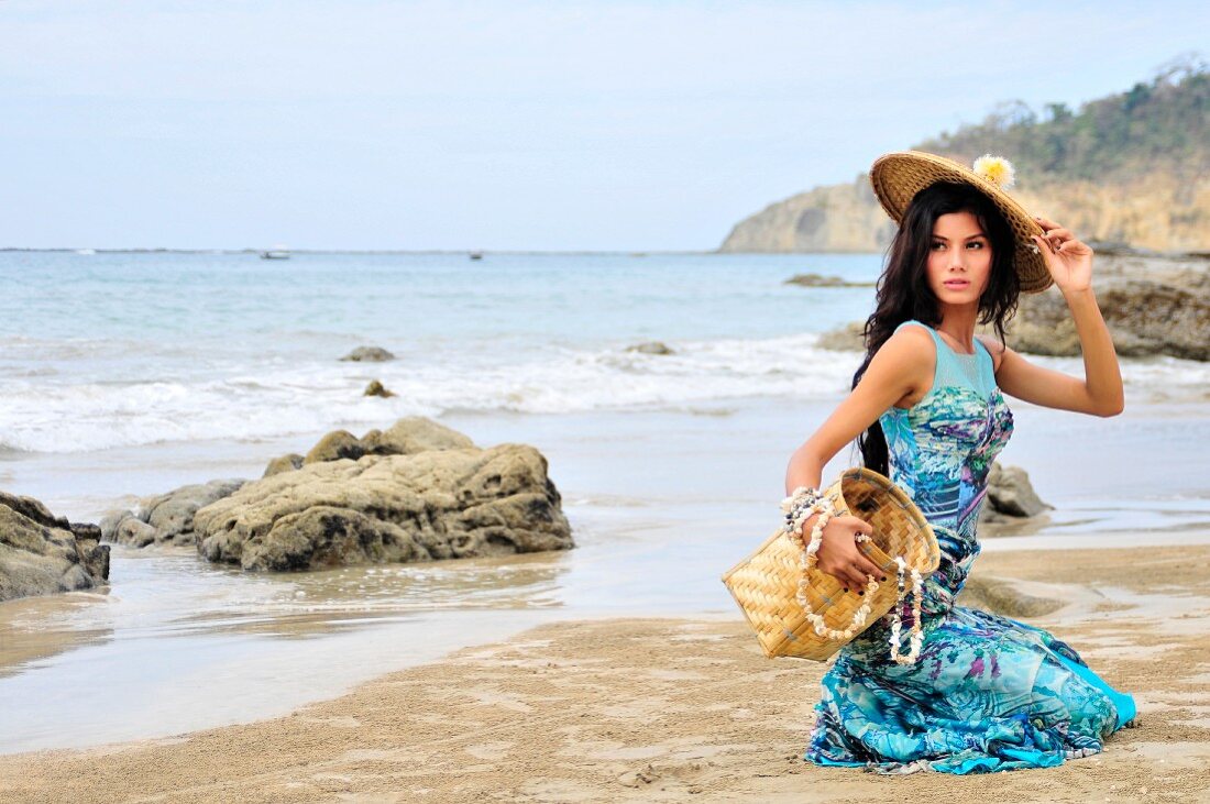 A woman kneeling on a beach wearing a turquoise patent dress and a straw hat