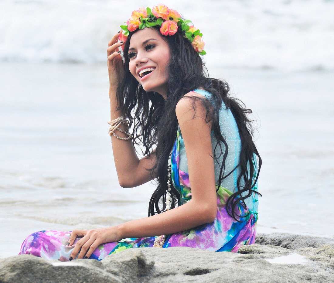 A woman sitting on a beach wearing brightly patterned stress and a wreath of flowers on her head