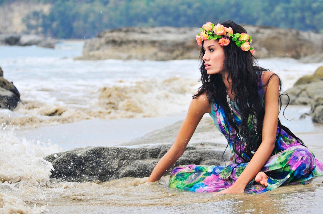 A woman sitting on a beach wearing brightly patterned stress and a wreath of flowers on her head