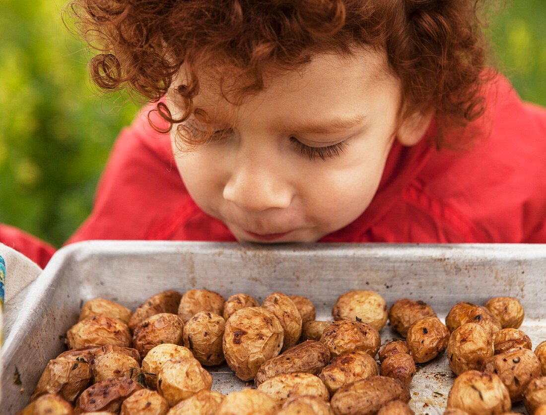 A red-haired boy sniffing at a tray of roast potatoes