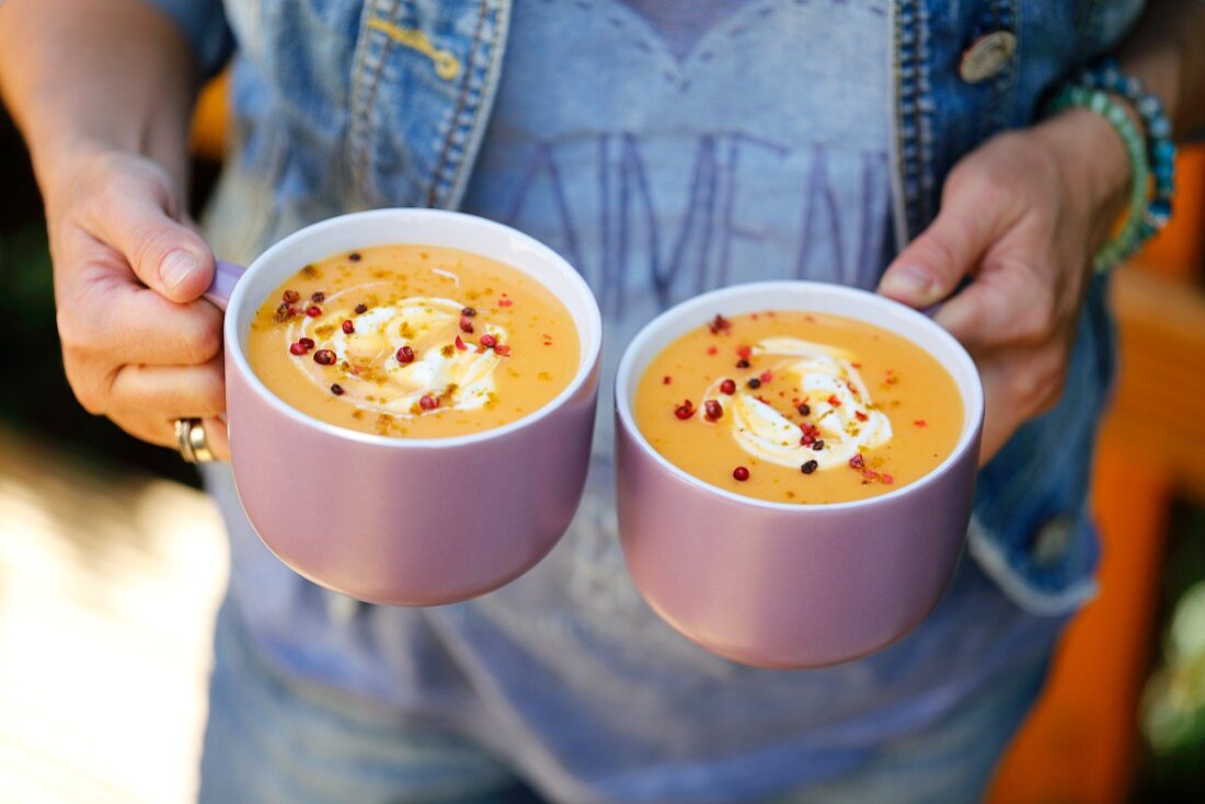 A woman holding two cups of cream of pumpkin soup with curry
