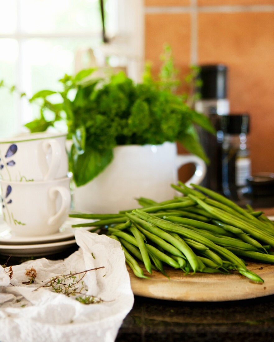 Green beans on a wooden plate in front of porcelain crockery with a herb bouquet