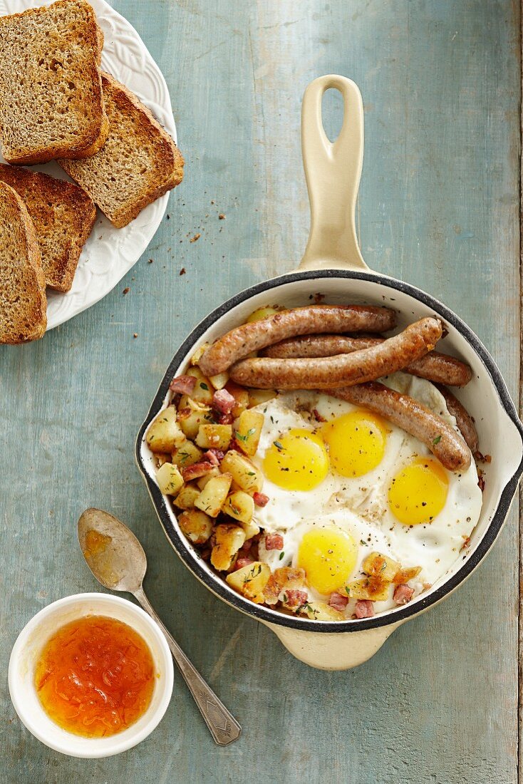 Frühstückseier mit Bratwürsten und Bratkartoffeln, dazu Vollkorntoast und Aprikosenmarmelade
