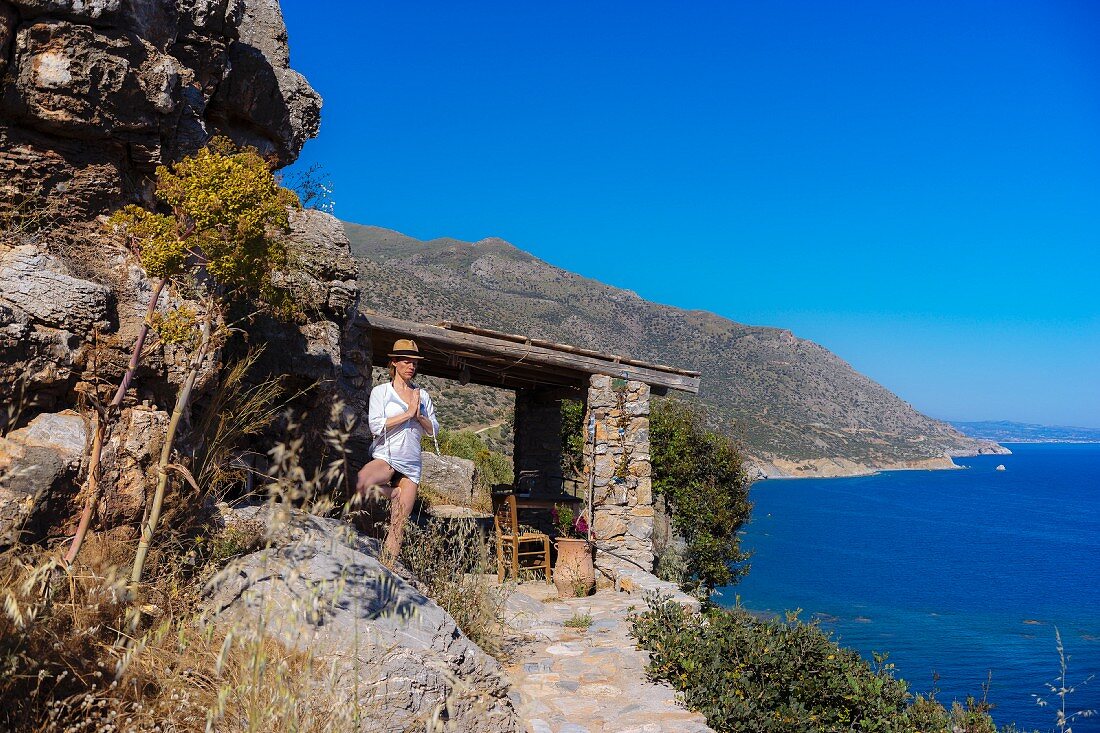 A woman practising yoga by a rustic stone house above a bay