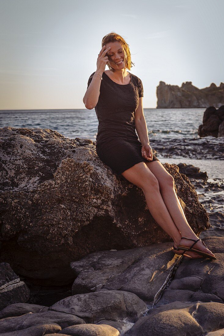 A woman wearing a black mini dress sitting on rocks by the sea