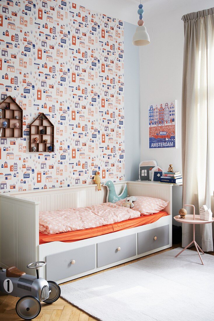 Grey and white wooden bed with storage drawers and orange fitted sheet against patterned wallpaper in child's bedroom