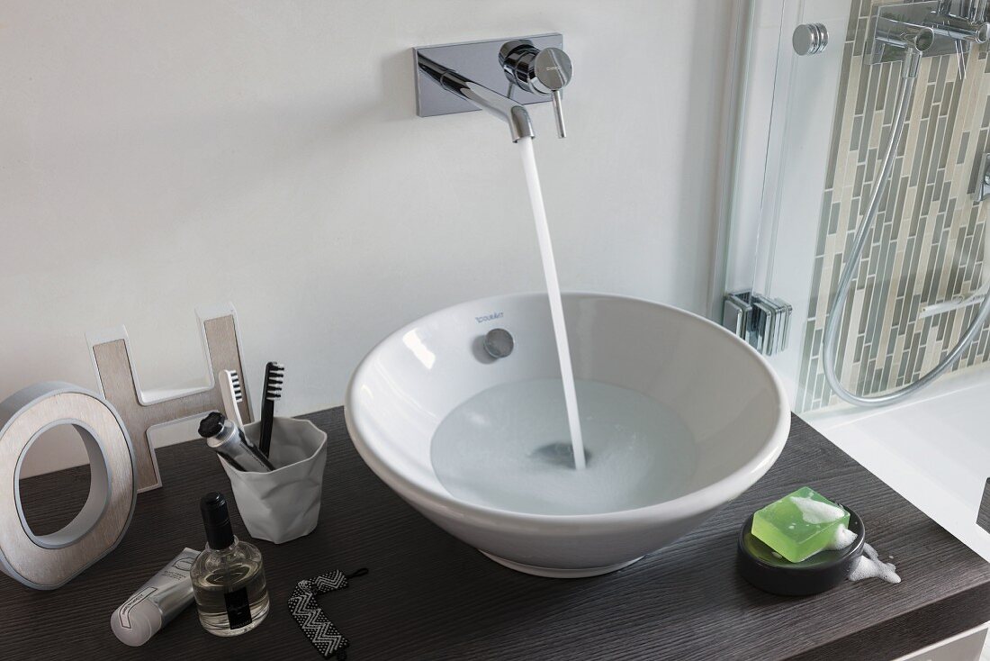 A wash stand with a bowl basin with water flowing into it from a wall mounted designer tap