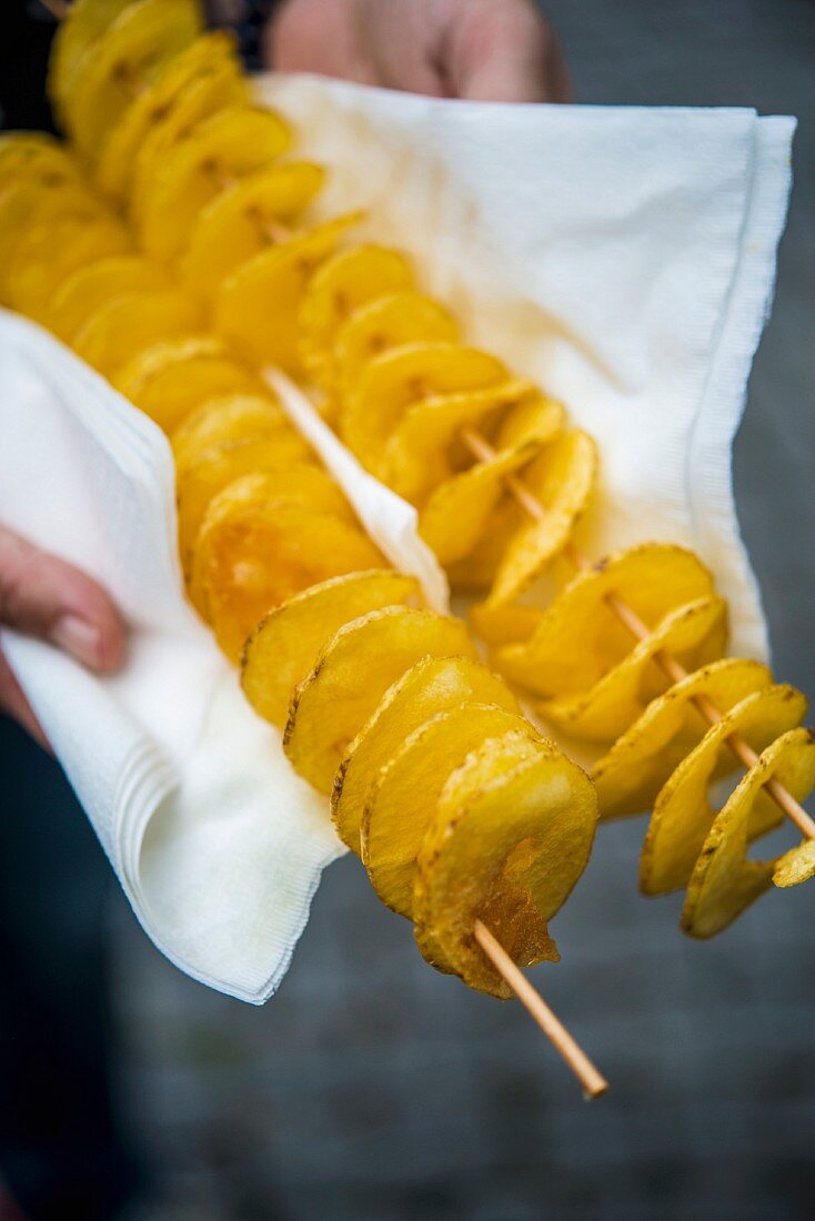 Hands holding potato spirals on wooden skewers in a napkin