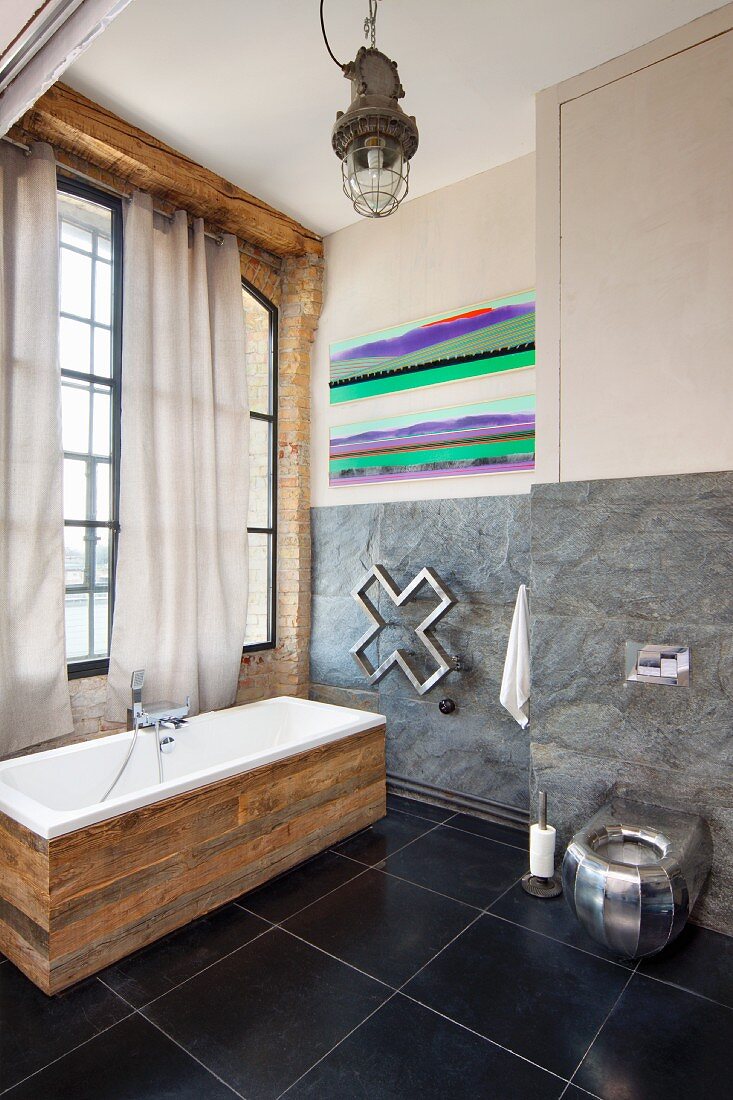 Free-standing bathtub with rustic wooden surround on black tiled floor in loft-style interior