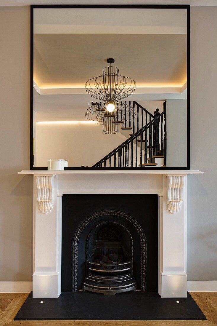 Mirror on white mantelpiece reflecting staircase and designer lamp in renovated interior