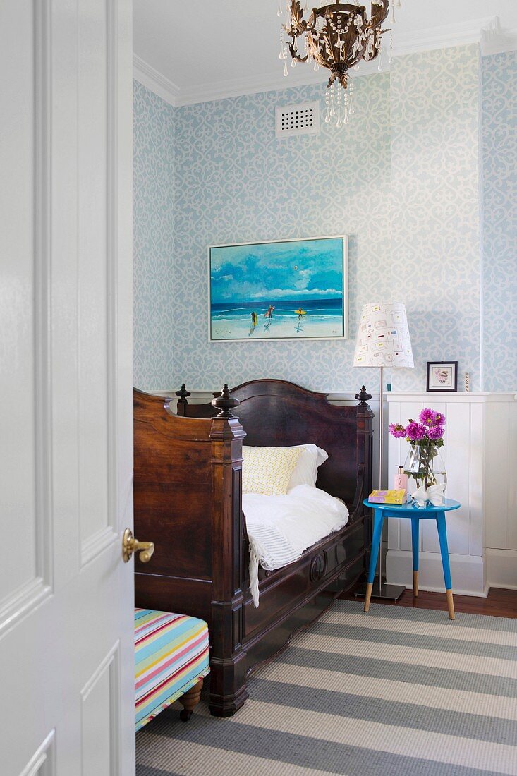 View through open room door of antique sleigh bed and blue bedside table in front of wood-paneled and wallpapered wall