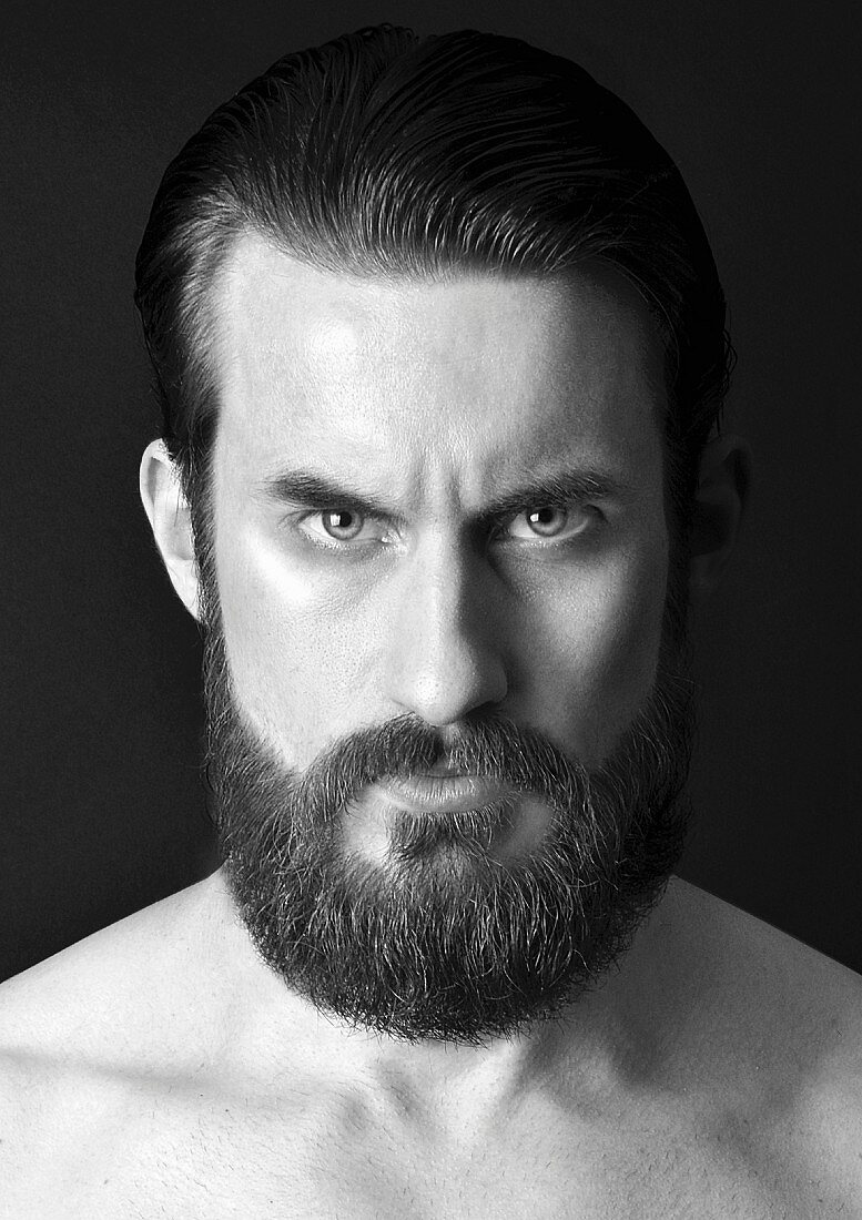 A fierce looking man with a full beard and his hair combed back (black-and-white shot)
