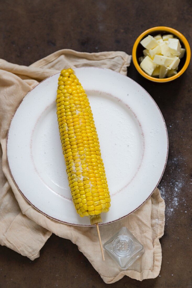 A cooked corn cob on a white plate with knobs of butter