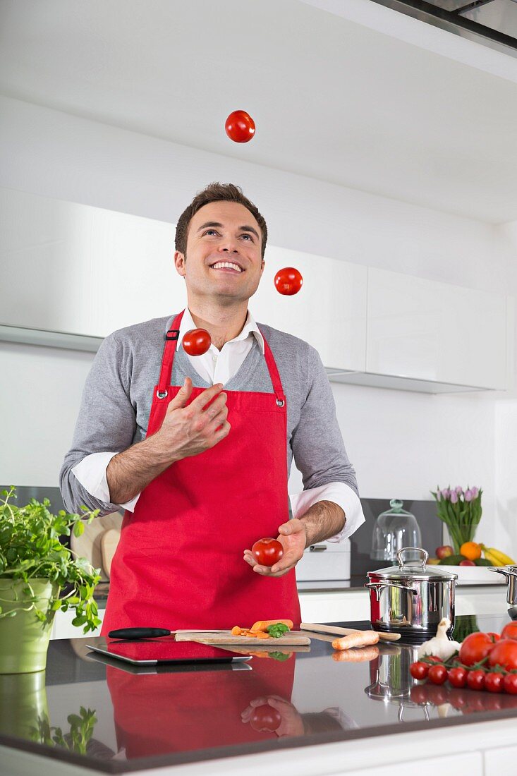 Mann mit Schürze jongliert lächelnd mit Tomaten in Küche