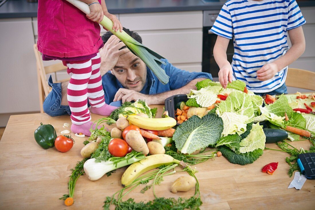 Verzweifelter Vater mit Kindern in Küche inmitten eines chaotischen Gemüsehaufens