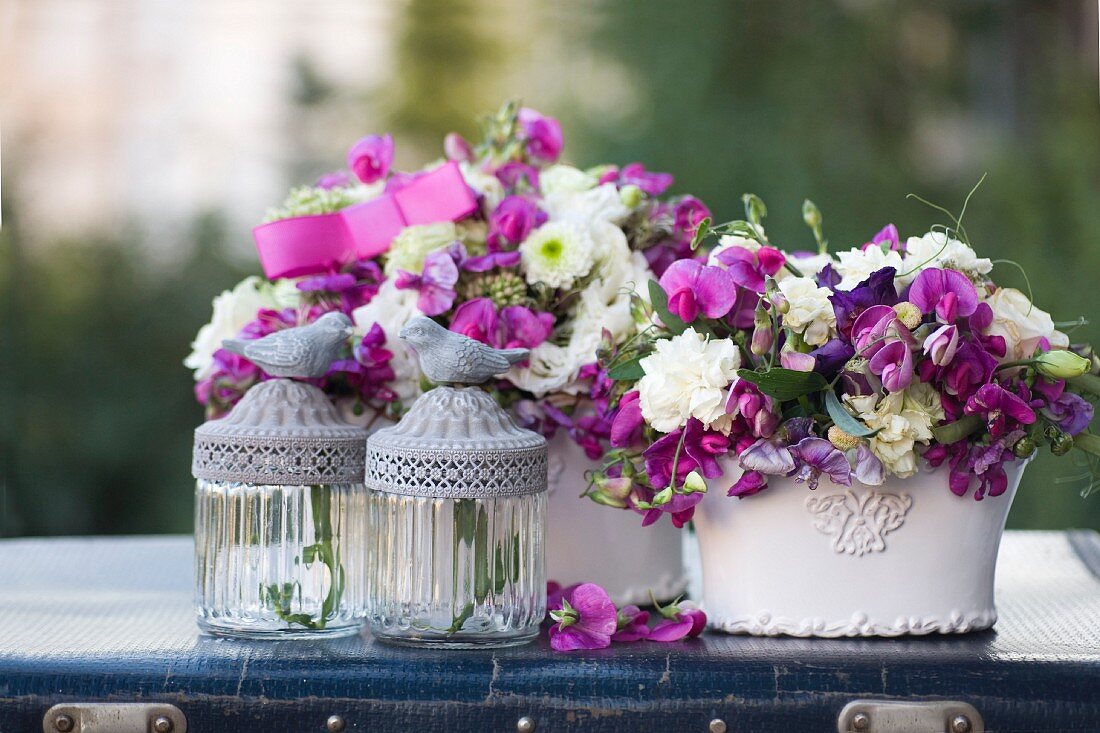 Two romantic flower arrangements and glass candle lanterns with bird figurines on lids on vintage suitcase