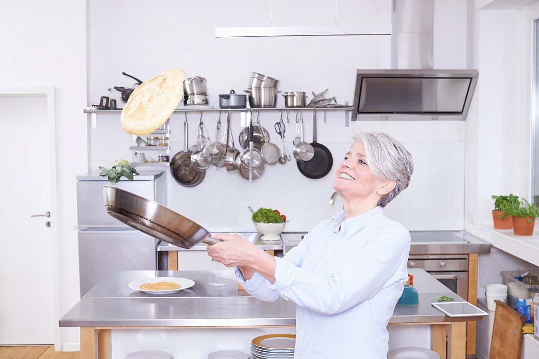 A middle aged woman in a kitchen tossing a pancake