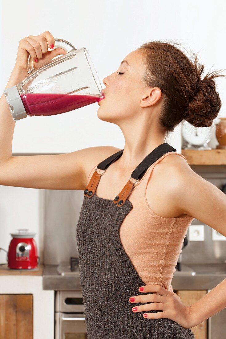Young woman wearing pink top and dungarees drinking red smoothie from mixer
