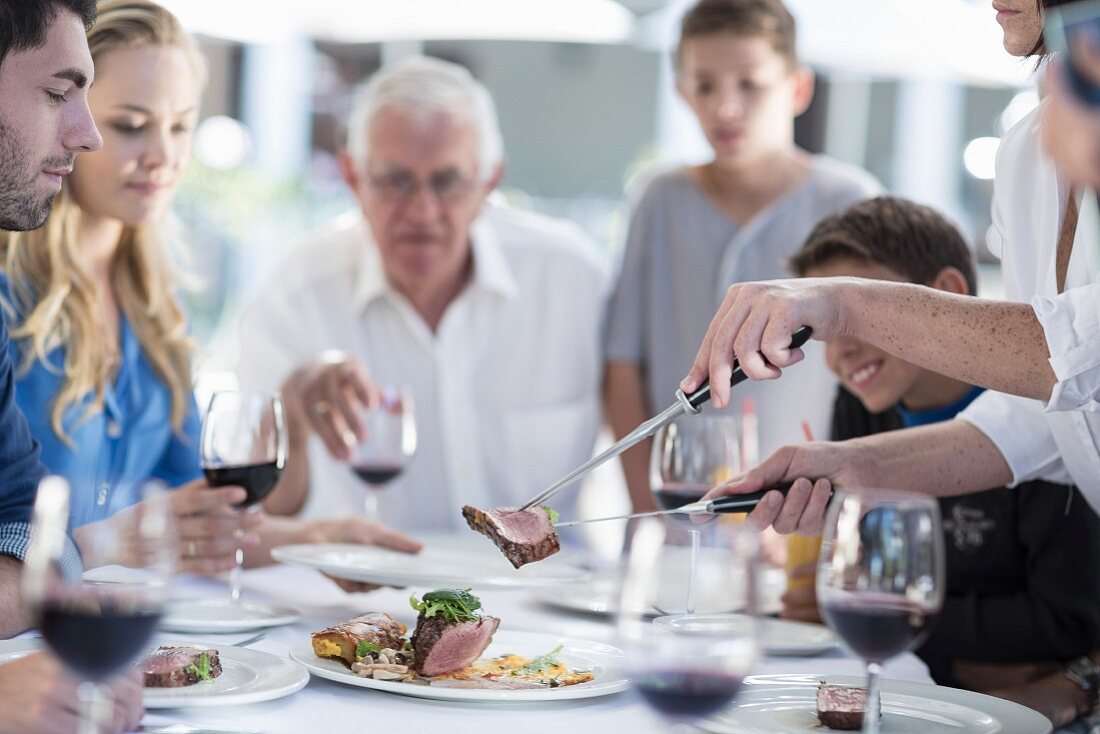 Familie beim gemeinsamen Essen am Tisch: Person schneidet & serviert Fleisch