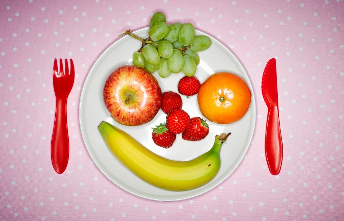A face made from fresh fruit on a plate with plastic cutlery (seen from above)