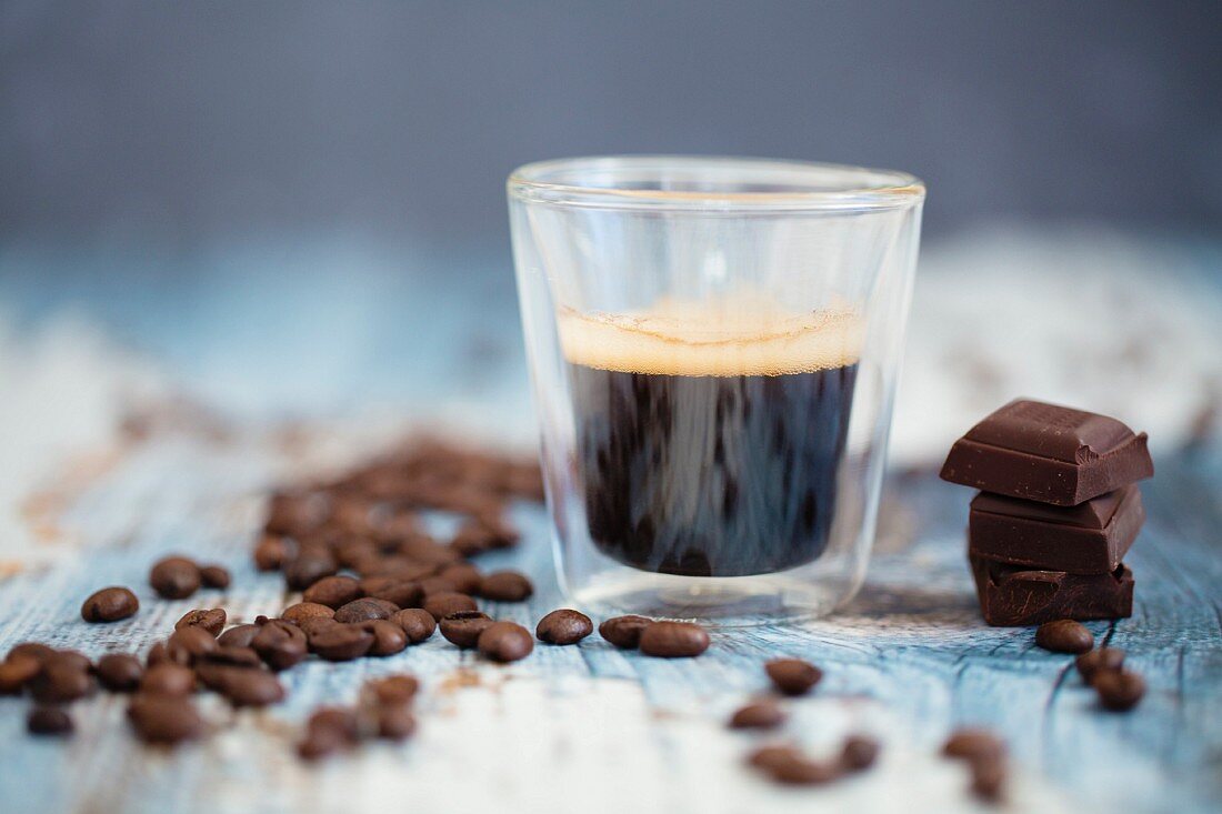 Espresso in Thermoglas daneben Kaffeebohnen & Schokoladenstücke