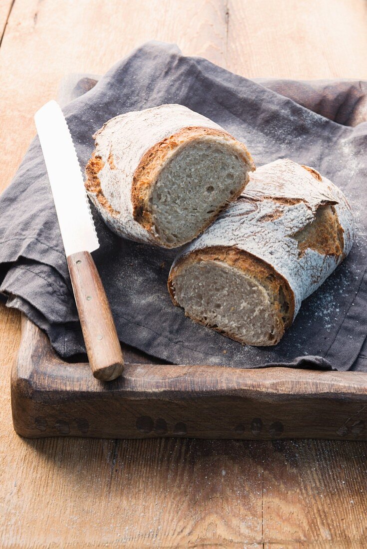 Two rustic loaves of bread on a linen cloth on a wooden tray with a bread knife