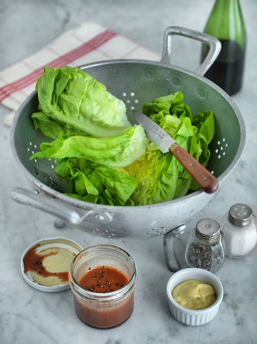 Lettuce in a colander with salad dressing, mustard, salt and pepper next to it