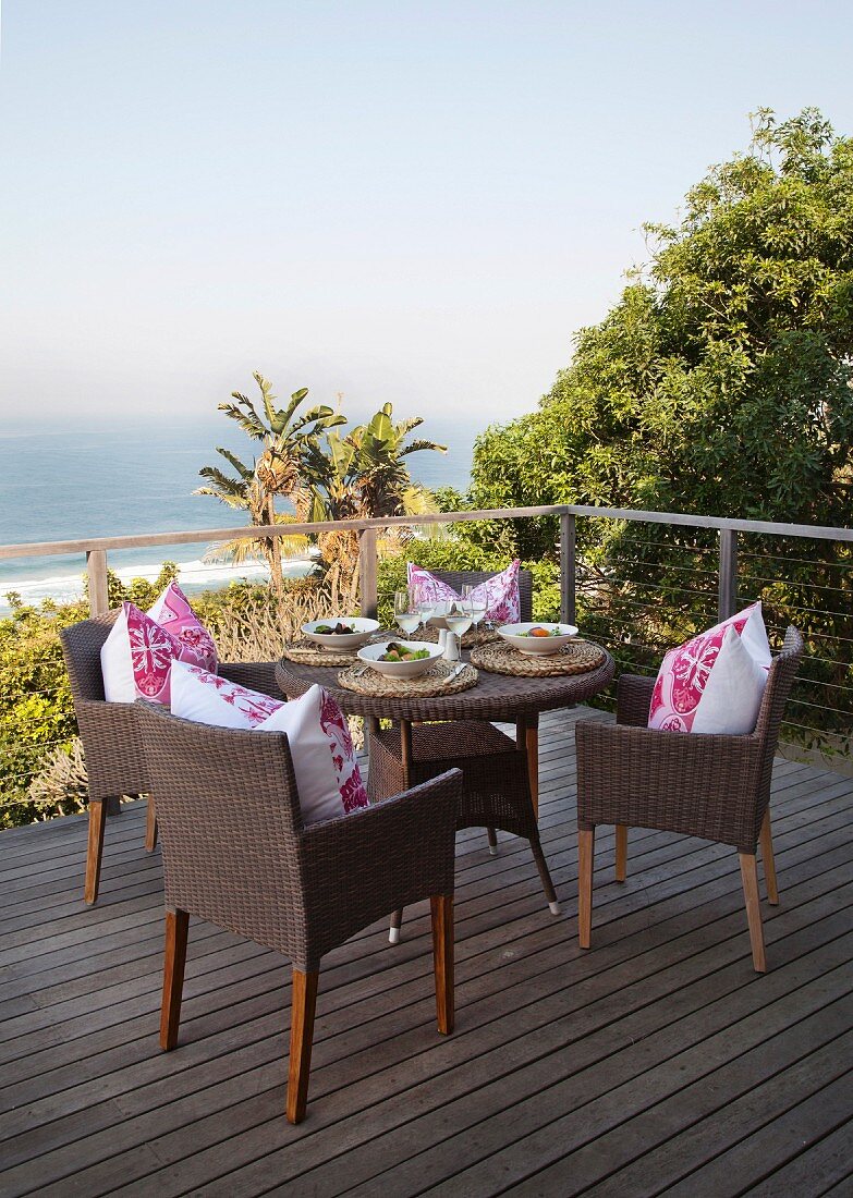 Holzterrasse mit gedecktem rundem Korbtisch und Blick über Palmen und Meer