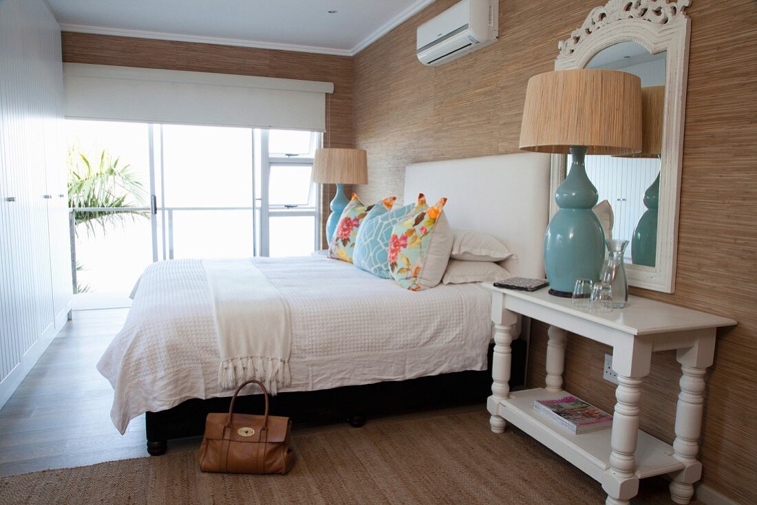 Bedroom with white fitted wardrobe, turquoise table lamp, scatter cushions on bed and brown wallpaper