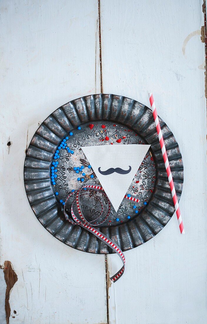 A ribbon, a straw, a paper moustache and sugar pearls on a metal plate