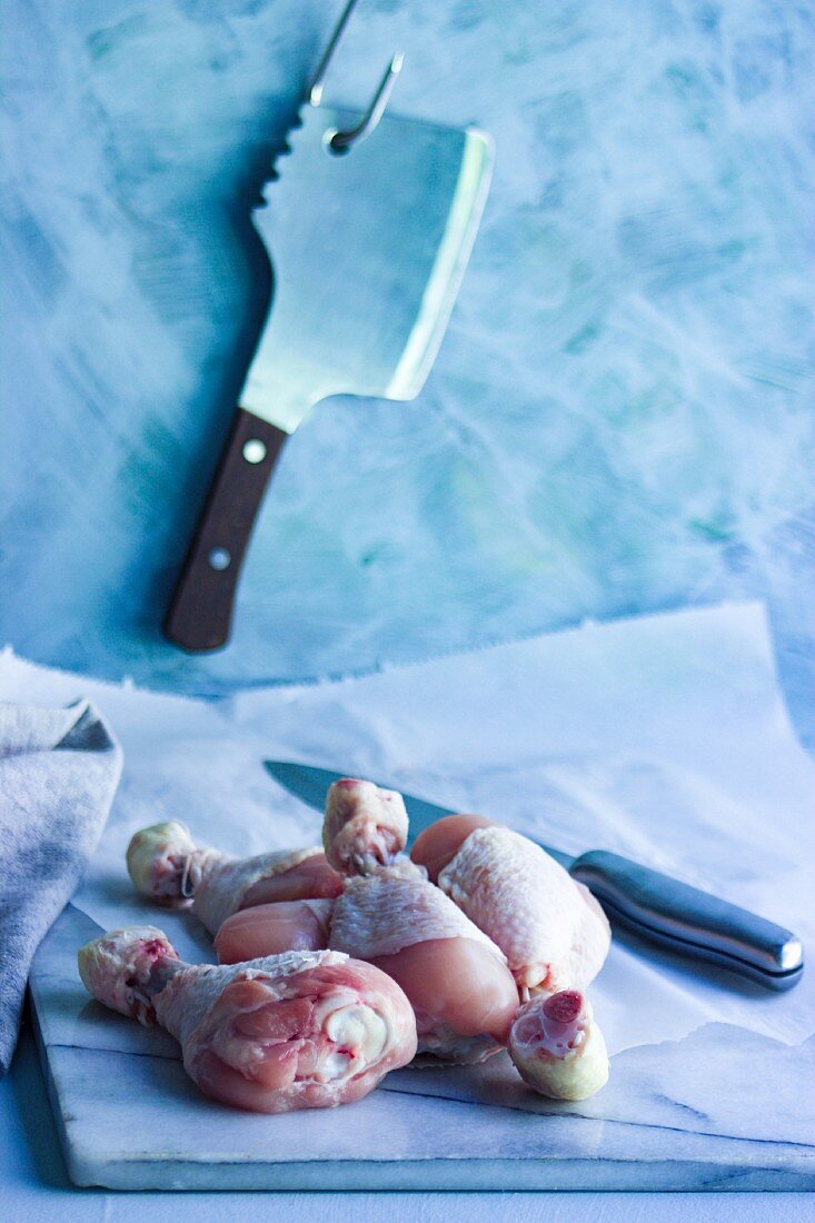 Raw chicken legs being prepared on a marble platter with a butcher's meat cleaver in the background