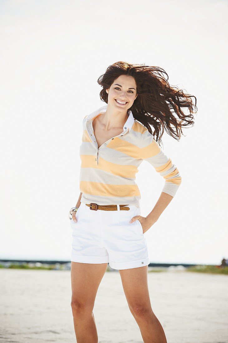 A brunette woman by a body of water wearing a long-sleeved T-shirt and a pair of shorts