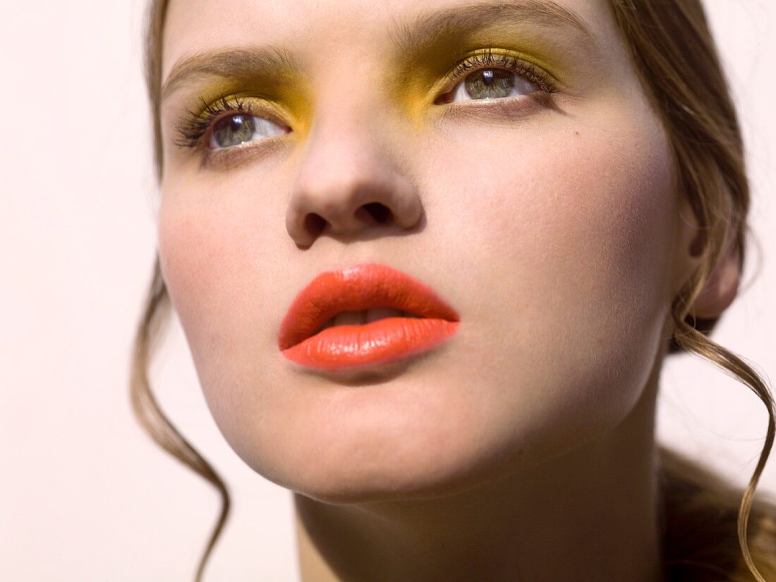A close-up portrait of a young woman wearing yellow eyeshadow and orange lipstick