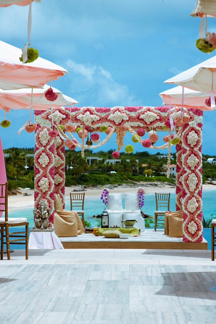 Indian outdoor wedding on platform; pergola decorated with flowers above chairs with white loose covers for bride and groom in front of sea coast
