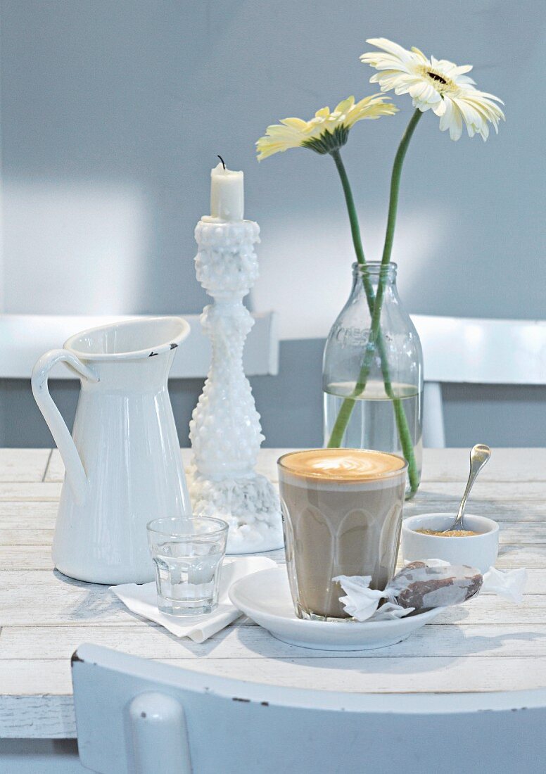 Cafe latte and sugar on a white wooden table with candle holder and a vase of flowers