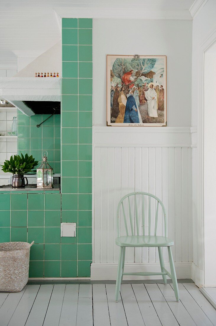 Pastellgrüner Stuhl vor weisser Holzverkleidung neben grün gefliestem Kochbereich in skandinavischer Küche